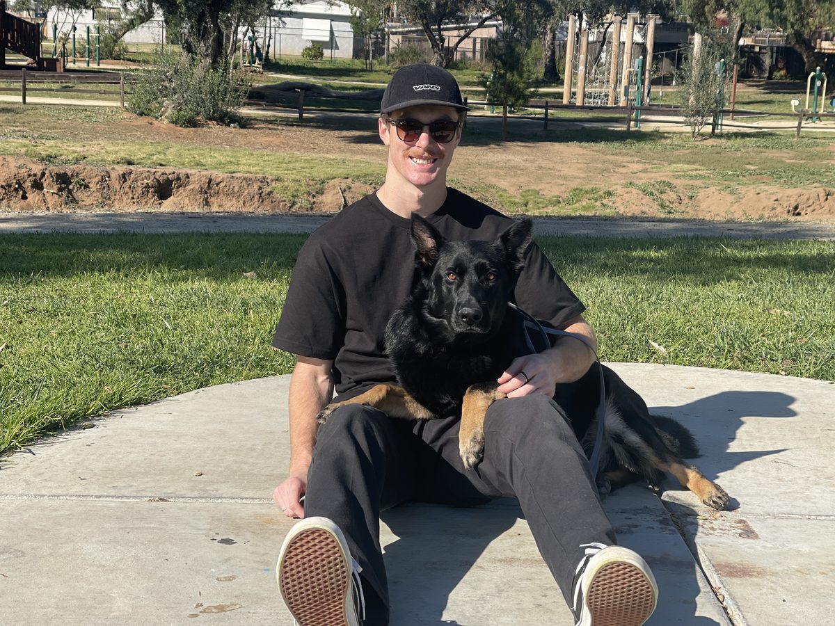 A trainer sitting on a path in a San Diego park with a calm German Shepherd across his lap